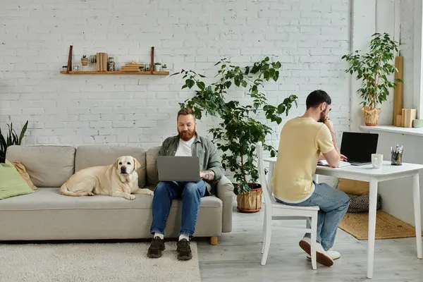 A bearded gay couple sits on a couch in their living room, working remotely beside their beloved labrador dog. — Stock Photo