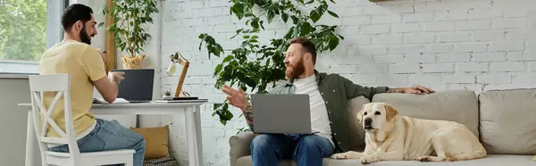 Una coppia gay barbuto seduto su un divano, lavorando su computer portatili, con il loro cane labrador nelle vicinanze in un ambiente accogliente soggiorno. — Foto stock