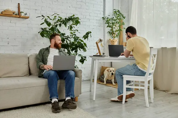 Un par de hombres están sentados encima de un sofá en una sala de estar, trabajando remotamente con un perro labrador cerca. - foto de stock