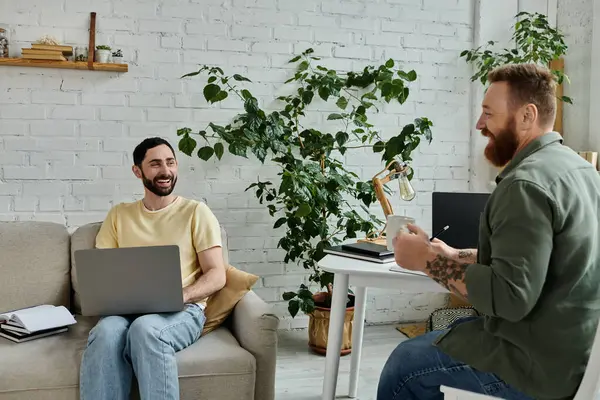 A bearded gay couple sits on a couch, one working on a laptop while the other looks on, near a labrador dog in a cozy living room. — Stock Photo