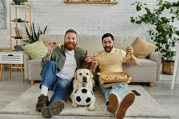 Двоє чоловіків, бородата гомосексуальна пара, сидять на дивані, їдять піцу і п'ють пиво. Лабрадорський собака приєднується до них у їх затишній вітальні. — стокове фото