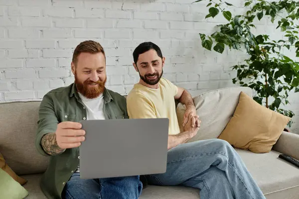 Бородата пара гей сидить на дивані, зосереджена на екрані ноутбука в їх затишній вітальні. — стокове фото
