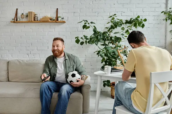 Un homme barbu dans un couple gay assis sur un canapé, tenant un ballon de football, profitant de temps de qualité dans leur salon. — Photo de stock