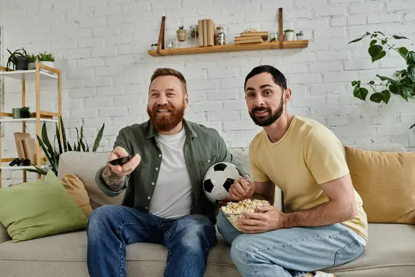 Two bearded men sit on a couch holding a soccer ball, enjoying quality time together in their cozy living room. — Stock Photo