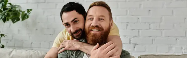 A bearded gay couple share a warm embrace while sitting together on a cozy couch in their living room. — Stock Photo
