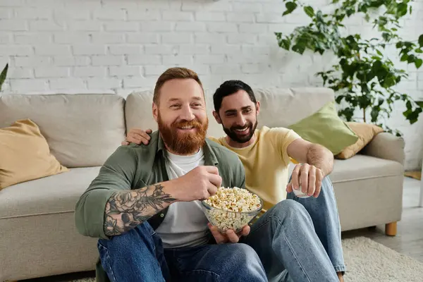 Dos hombres, una pareja gay barbuda, se sientan en un sofá, disfrutando de palomitas de maíz mientras pasan tiempo juntos en la sala de estar. - foto de stock