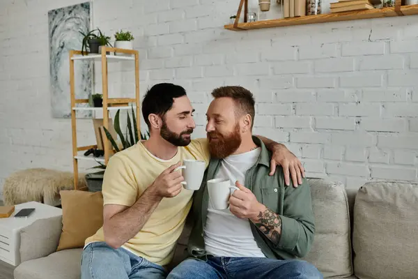 Um casal gay barbudo relaxa juntos, sentados em cima de um sofá em sua sala de estar, desfrutando de tempo de qualidade. — Fotografia de Stock