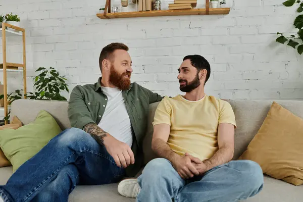 Bearded gay couple sitting on a couch, engaged in a heartfelt conversation in a cozy living room setting. — Stock Photo