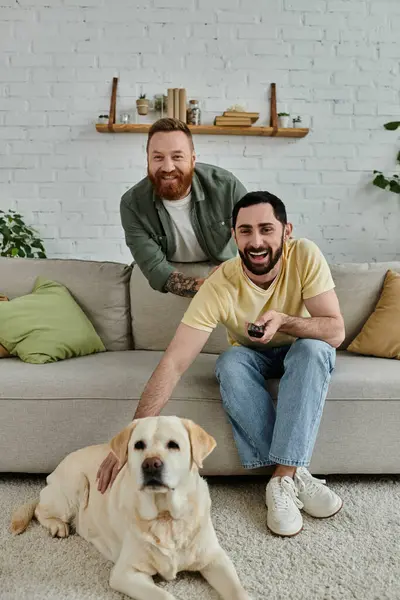 Um homem com barba sentado em um sofá ao lado de um Labrador retriever em uma acolhedora sala de estar. — Fotografia de Stock