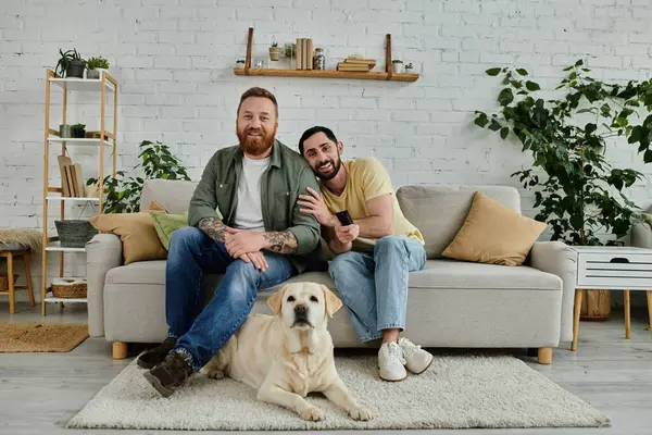Two bearded men sitting on a couch, enjoying quality time together with their Labrador in a cozy living room setting. — Stock Photo