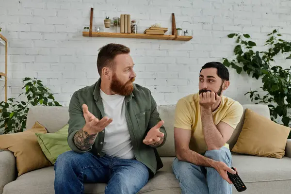 Bearded gay couple engaged in lively discussion, seated on couch in cozy living room — Stock Photo