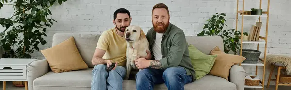 Two men, one bearded, relaxing on a couch with their Labrador in a cozy living room setting. — Stock Photo