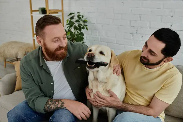 Bearded gay couple sitting on a couch, joyfully holding and petting their Labrador in a cozy living room setting. — Stock Photo