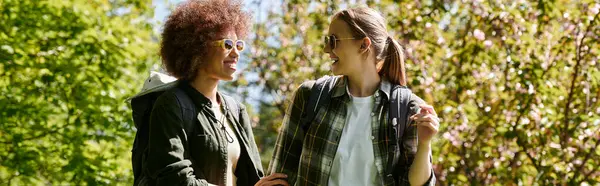 Two young women, dressed in casual outdoor wear, hike through a forest on a sunny day. — Stock Photo