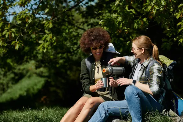 Two young lesbian women enjoying a break during their hike, one pouring tea from a thermos into a mug for her partner. — Stock Photo