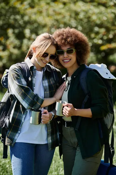 Two young women, one with curly hair, hike through the woods together, carrying backpacks and enjoying coffee. — Stock Photo