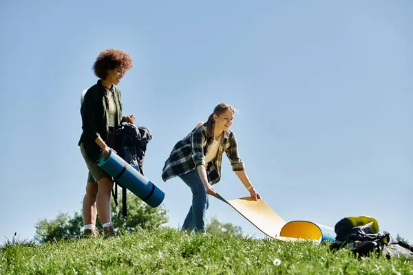 Two young women prepare for a camping trip in the wilderness, rolling out a sleeping pad on the grassy ground. — Stock Photo