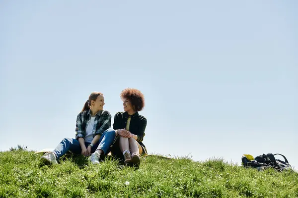 Dos mujeres jóvenes, una de cabello castaño y otra de pelo oscuro rizado, están sentadas en una colina cubierta de hierba, disfrutando de su caminata.. - foto de stock
