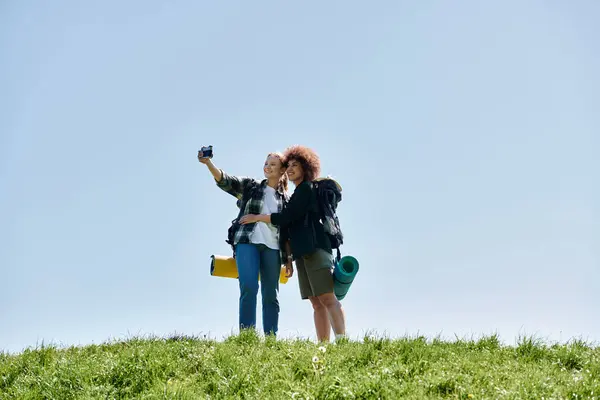 Two young women, a lesbian couple, hike together in the wilderness and take a selfie on a grassy hill. — Stock Photo