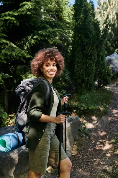 Una mujer sonríe mientras camina a través de un exuberante bosque verde, su mochila y bastones de senderismo a su lado. — Stock Photo