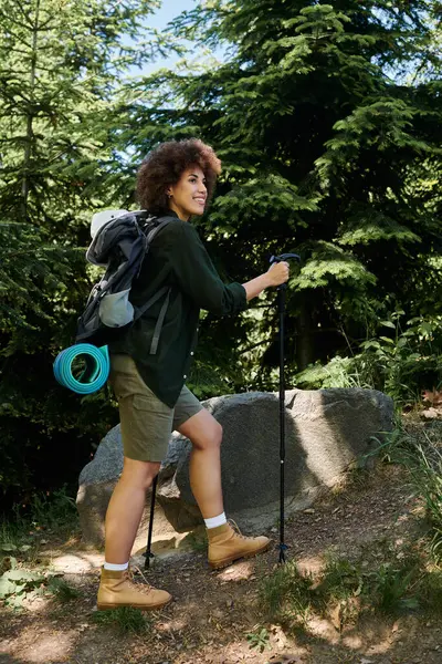 Una giovane donna cammina attraverso una foresta estiva, indossando uno zaino e utilizzando bastoncini da trekking. — Foto stock