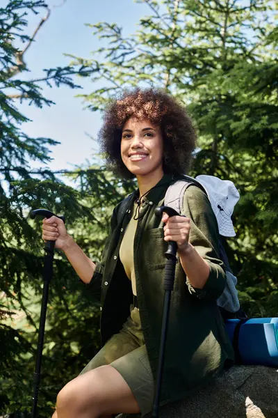 A young woman smiles while hiking in the woods during the summer. She is holding two trekking poles and wearing a backpack. — Stock Photo