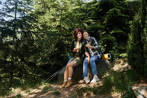 A young lesbian couple enjoys a snack break on a rocky trail during a hike through a lush forest. — Stock Photo