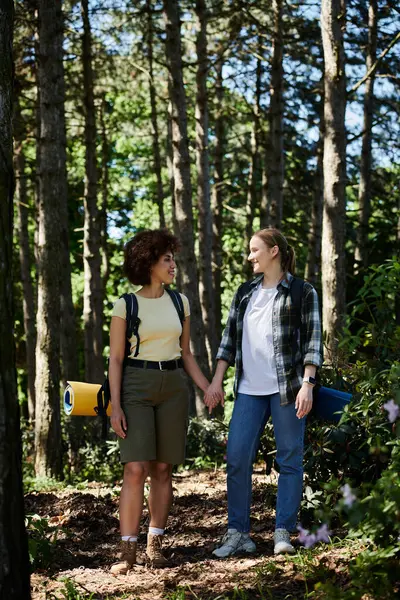 A young lesbian couple hikes through a forest, hand in hand, enjoying a sunny day. — Stock Photo