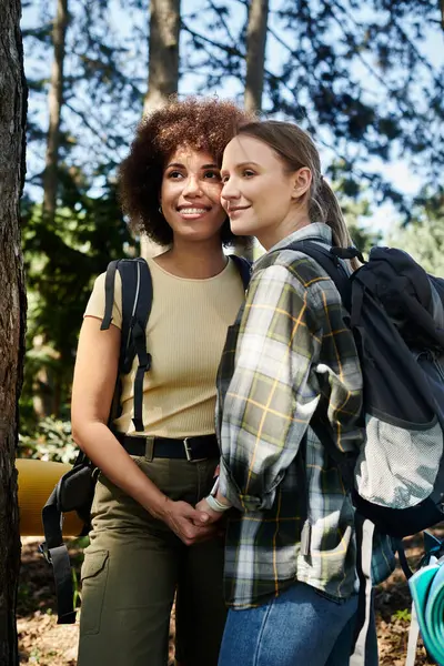 Two young women, one with curly brown hair and the other with straight blonde hair, hike through a wooded area, smiling and holding hands. — Stock Photo