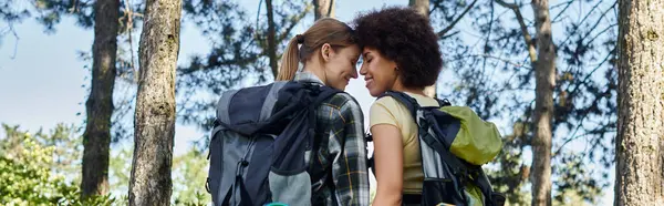 Two young women, one with blonde hair and one with curly brown hair, are hiking in the woods. They are wearing backpacks and looking at each other lovingly. — Stock Photo