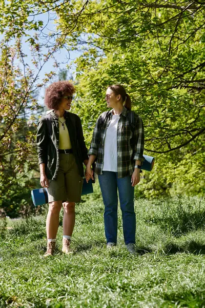 Two young women, one with short, curly hair and the other with long, straight hair, hike through a forest, their smiles reflecting the joy of their time together. — Stock Photo