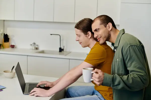 A young gay couple enjoys a casual morning in their modern kitchen, one partner working on a laptop while the other embraces him from behind. — Stock Photo