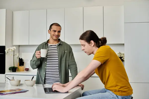 A young gay couple relaxes in their modern apartment, one holding a mug of coffee while the other works on a laptop. — Stock Photo