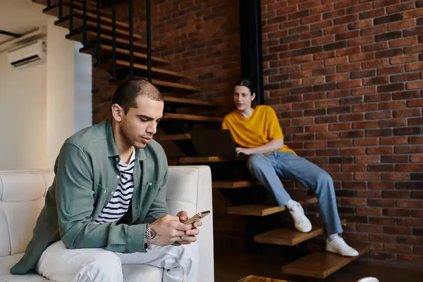 A young gay couple relaxes in their modern apartment, one man on the couch looking at his phone, the other on the stairs using a laptop. — Stock Photo