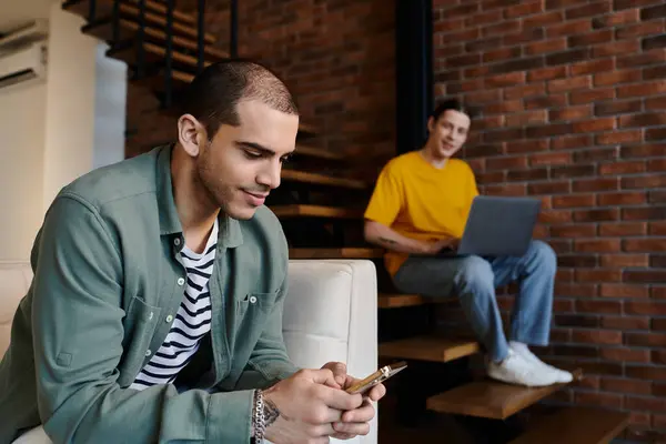 A young gay couple relaxes in their modern apartment, one scrolling through his phone while the other works on a laptop. — Stock Photo