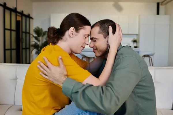 A young gay couple in casual clothing embraces on a white couch in their modern apartment. — Stock Photo