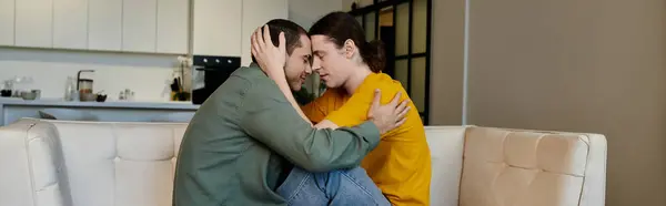 A young gay couple sits on a white couch in a modern apartment, their arms wrapped around each other. — Stock Photo