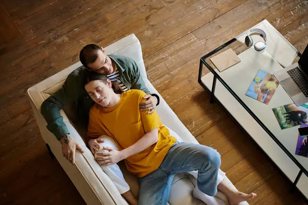 A young gay couple relaxes on a white couch in a modern apartment, one man resting his head on the others shoulder. — Stock Photo