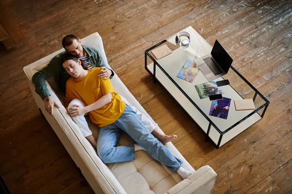 A young gay couple relaxes on a white couch in a modern apartment, showcasing their love and comfort in their shared space. — Stock Photo