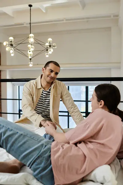 A young gay couple relaxes on a bed in a modern apartment. The man on the bed is holding the hand of the man standing above him. — Stock Photo