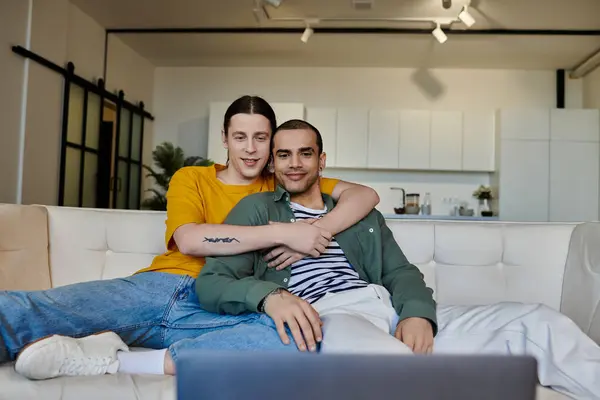 A young gay couple cuddles on a white couch in their modern apartment. — Stock Photo