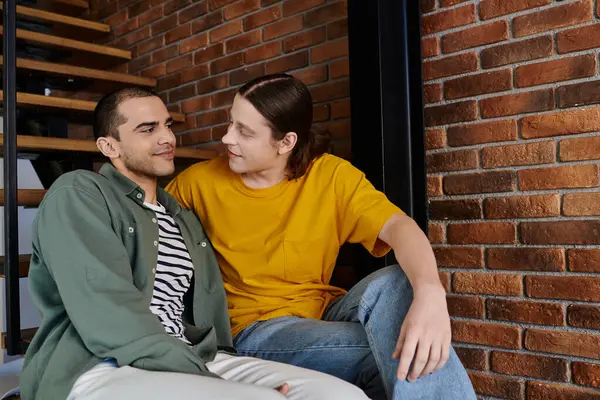 A young gay couple sits together in a modern apartment, their eyes locked in a tender moment of affection. — Stock Photo