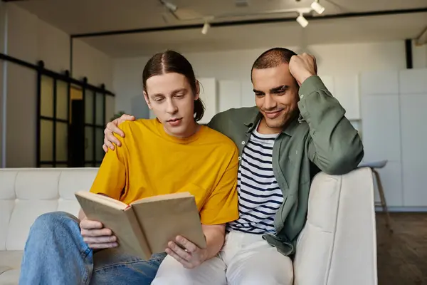 A young gay couple relaxes on a couch in their apartment, sharing a book together. — Stock Photo