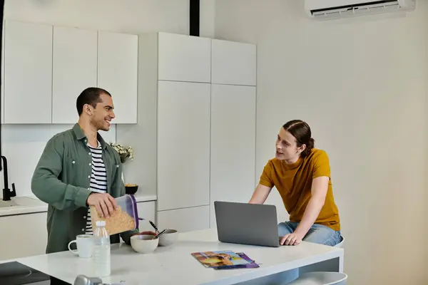 Un jeune couple gay profite d'une matinée décontractée dans leur appartement moderne, l'un préparant le petit déjeuner tandis que l'autre travaille sur un ordinateur portable. — Photo de stock