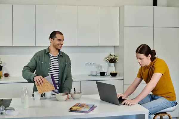 A young gay couple spends a relaxed morning in their modern apartment. One is pouring cereal while the other works on a laptop. — Stock Photo