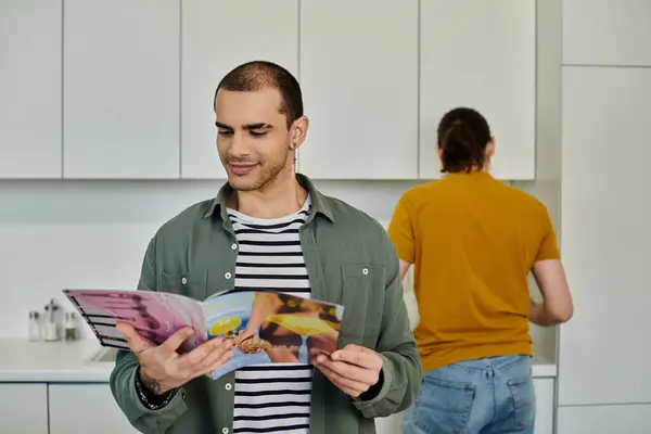 A young gay couple enjoys a cozy moment in their modern apartment, one man reading magazine while the other stands in the background. — Stock Photo