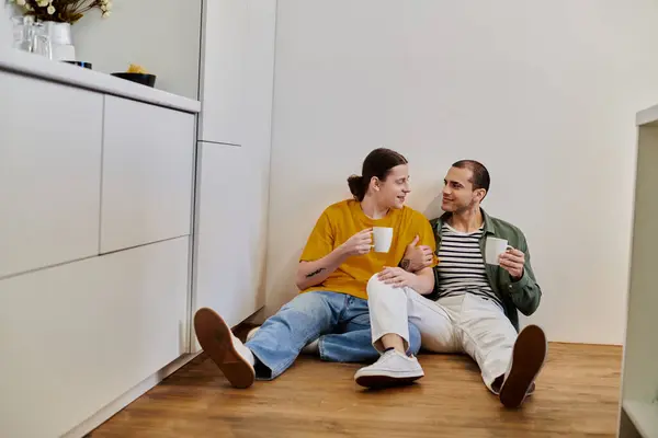 Un jeune couple gay s'assoit sur le sol de leur cuisine moderne, profitant d'une tasse de café et de l'autre compagnie. — Photo de stock