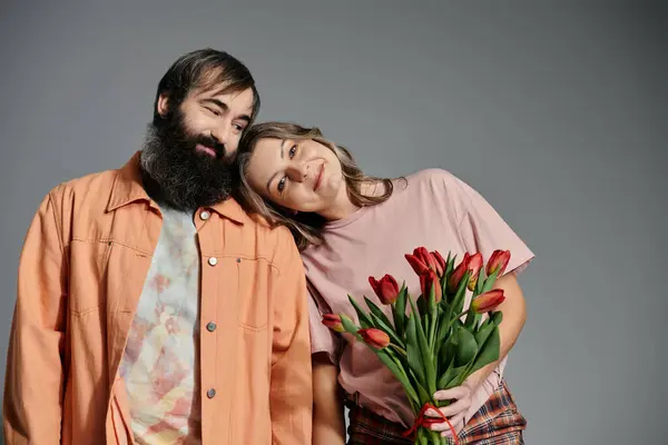 Um casal amoroso em trajes elegantes posa junto com um pano de fundo cinza, com a mulher segurando um buquê de tulipas vermelhas. — Fotografia de Stock