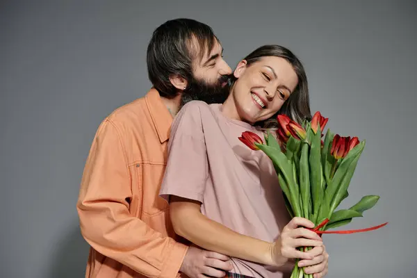 A couple in stylish attire embraces against a grey background, the woman holding a bouquet of red tulips. — Stock Photo
