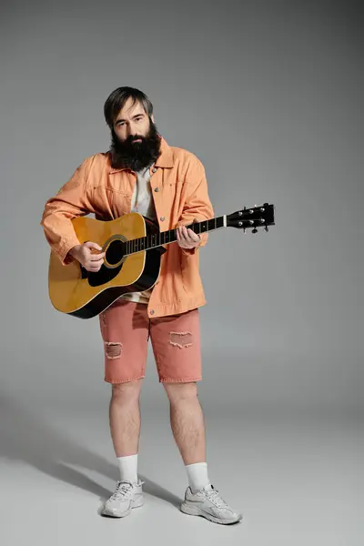 A man in a peach jacket and shorts plays a guitar while standing against a gray background. — Stock Photo